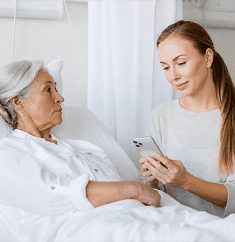 Young caregiver using smartphone to assist elderly woman in hospital bed.